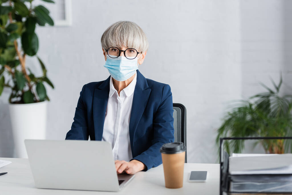 leader del team di mezza età in occhiali e maschera medica utilizzando laptop vicino smartphone e tazza di carta sulla scrivania - Foto, immagini