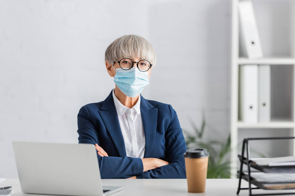 middle aged team leader in glasses and medical mask sitting with crossed arms near laptop and paper cup on desk - Photo, Image