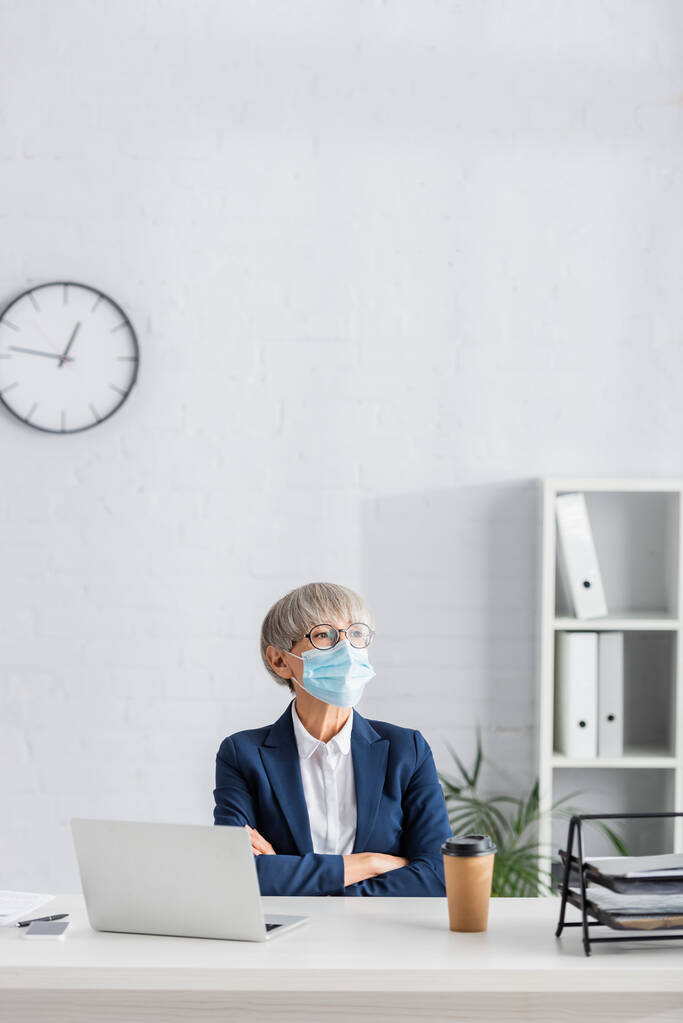 dojrzały lider zespołu w okularach i masce medycznej siedzi ze skrzyżowanymi ramionami w pobliżu laptopa i kubka papieru na biurku - Zdjęcie, obraz