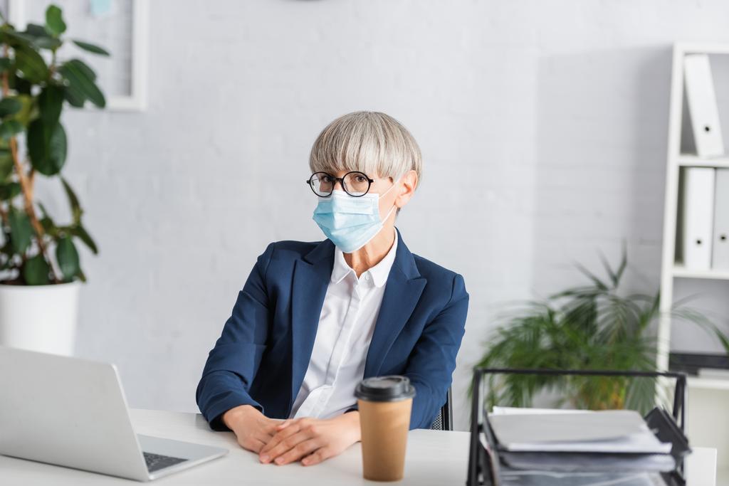 mature team leader in glasses and medical mask looking at camera near laptop and paper cup on desk - Photo, Image