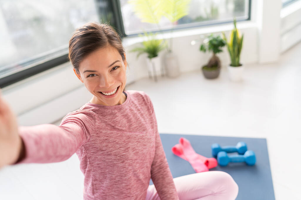 Щаслива дівчина з Азії, яка посміхається тренінги для тіла на маті у вітальні, фотографує мобільний під час тренування. - Фото, зображення