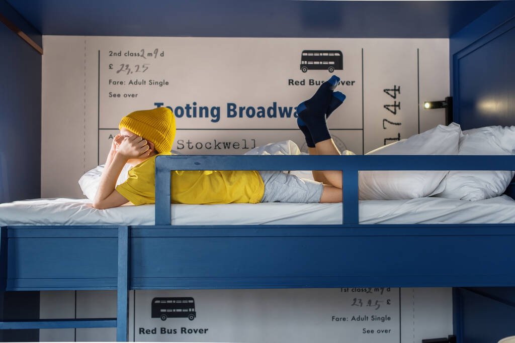 Chlapec ve žlutém tričku a klobouku tráví čas ležením v županu na modré dvoupatrové palandě doma. - Fotografie, Obrázek
