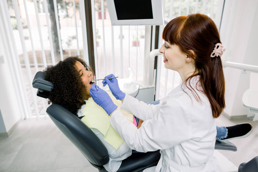 Dentist and kid at modern dental clinic. Young smiling caucasian dentist in white uniform and gloves, making examination of her small patient, little mixed raced curly girl, to prevent caries - Foto, imagen