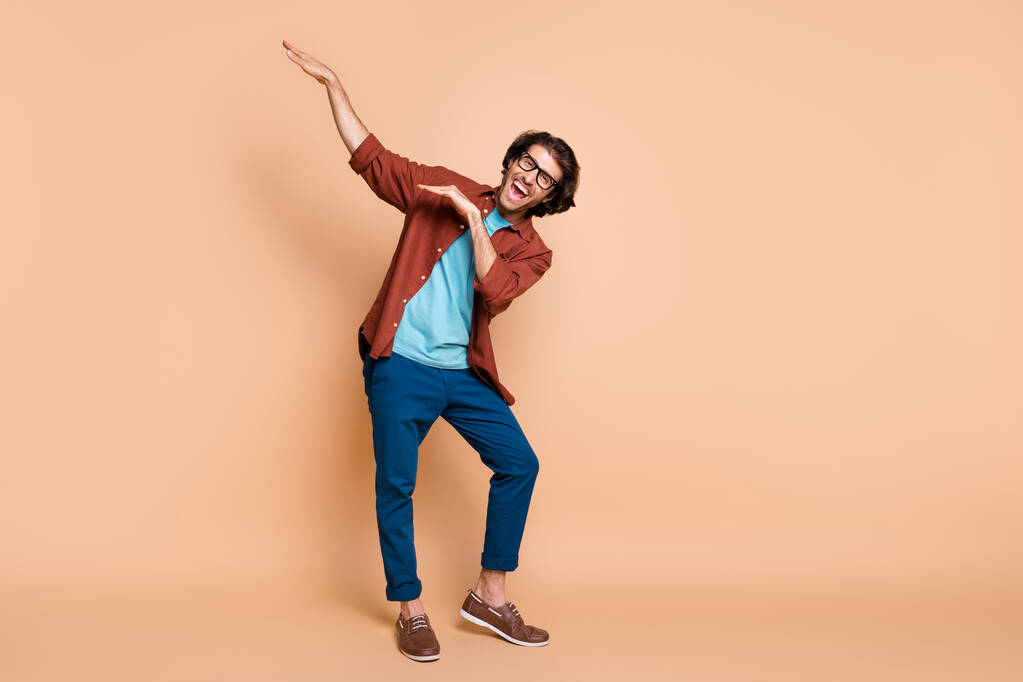 Full length photo of guy dance raise hands have fun wear spectacles brown t-shirt trousers shoes isolated beige color background - Photo, Image