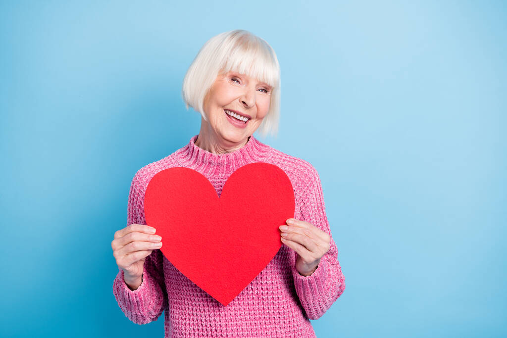 Foto retrato de una linda anciana sosteniendo una postal de corazón rojo en dos manos aisladas sobre fondo de color azul pastel - Foto, Imagen