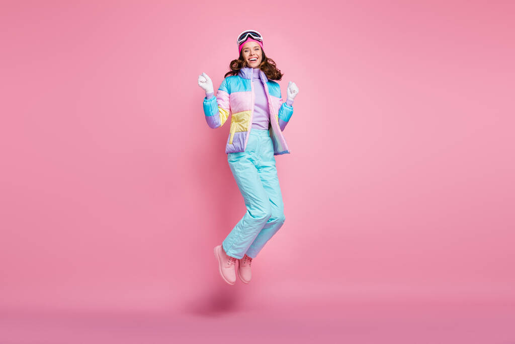 Foto de longitud completa de la mujer afortunada esquiadora vestida coloridos anteojos cortavientos puños ascendentes saltando aislado color rosa fondo - Foto, imagen