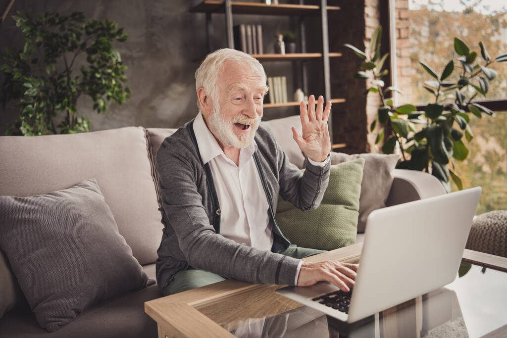 Foto di divertente pensionato indossare cardigan grigio seduto cercando gadget moderno hanno videochiamata all'interno casa piatta - Foto, immagini