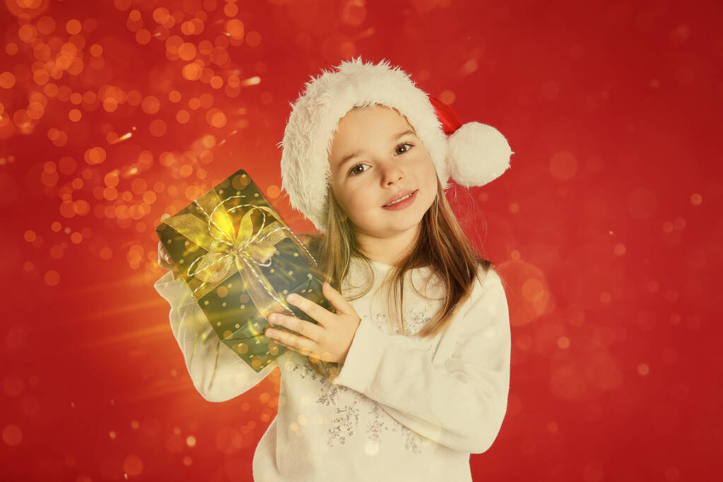Niedliches Kind in Weihnachtsmütze mit Weihnachtsgeschenk auf rotem Hintergrund - Foto, Bild