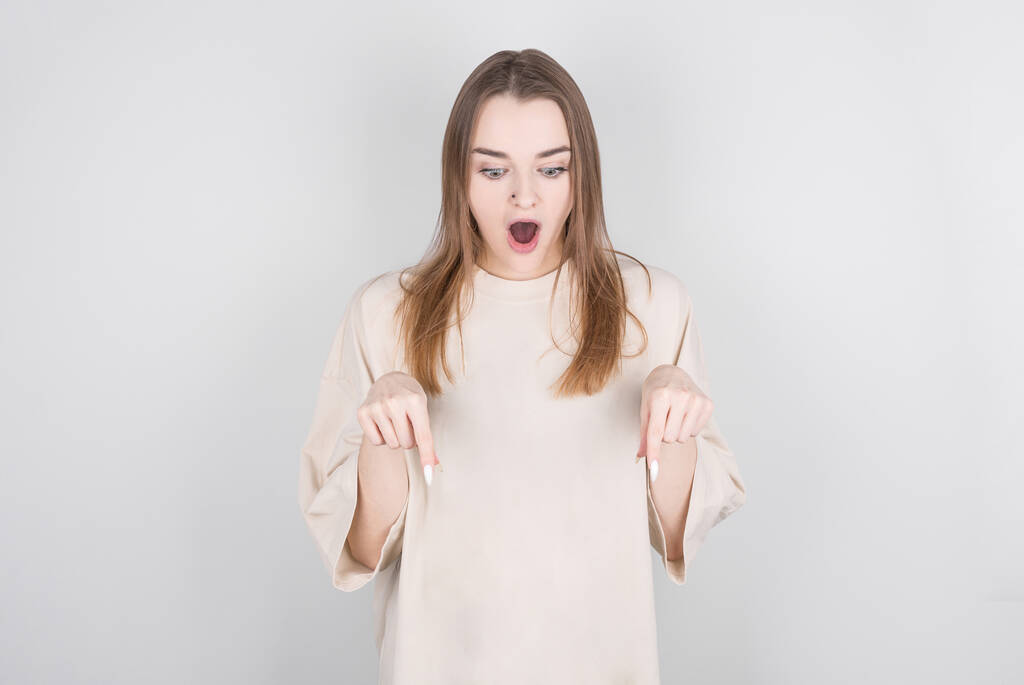 Photo of surprised brunette woman points on floor, has shocked facial expression, wears white t shirt against white background. Wow concept - Photo, Image