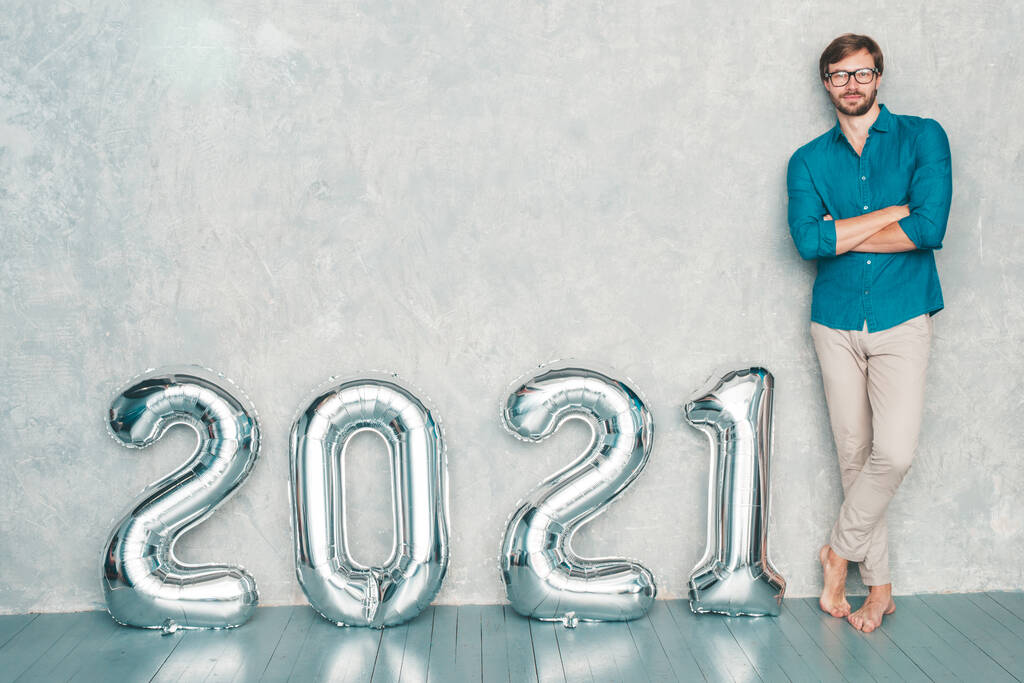 笑顔ハンサムな男が壁の近くにポーズの肖像画。シルバー2021風船の近くに立つセクシーなひげを生やした男性。新年明けましておめでとうございます。金属番号2021 - 写真・画像