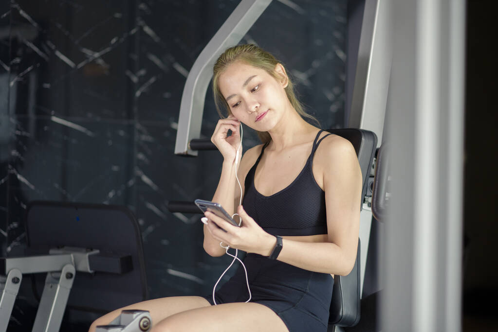 Μια όμορφη χαμογελαστή γυναίκα σε ακουστικά χρησιμοποιώντας ένα κοινωνικό δίκτυο και ακούγοντας μουσική στο γυμναστήριο, Fitness και την έννοια της τεχνολογίας - Φωτογραφία, εικόνα