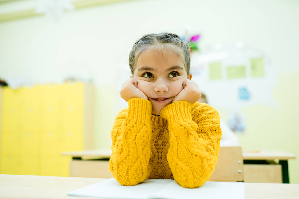 Egy sárga kötött pulóveres kisgyerek ül az iskolaasztalnál könyökével a kezén. - Fotó, kép
