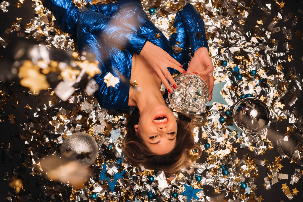 una mujer con un vestido de lentejuelas azules sonríe y yace en el suelo bajo un confeti multicolor que cae. - Foto, imagen