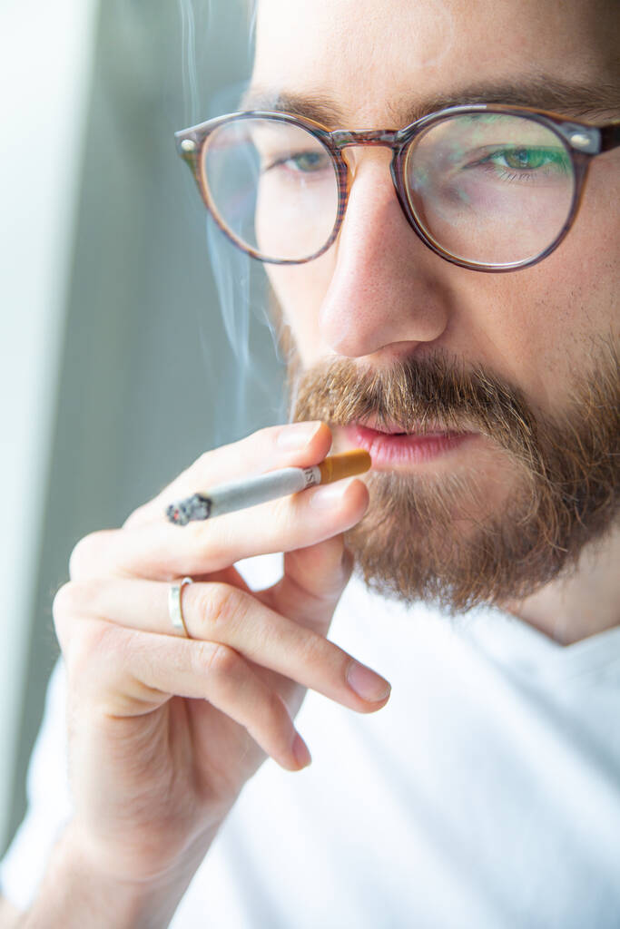 Молодой взрослый, бородатый мужчина в белой футболке курит сигарету крупным планом. Курение перерыв в течение рабочего дня, на открытом воздухе. Никотиновая зависимость. Карантин на дому. Депрессия.. - Фото, изображение