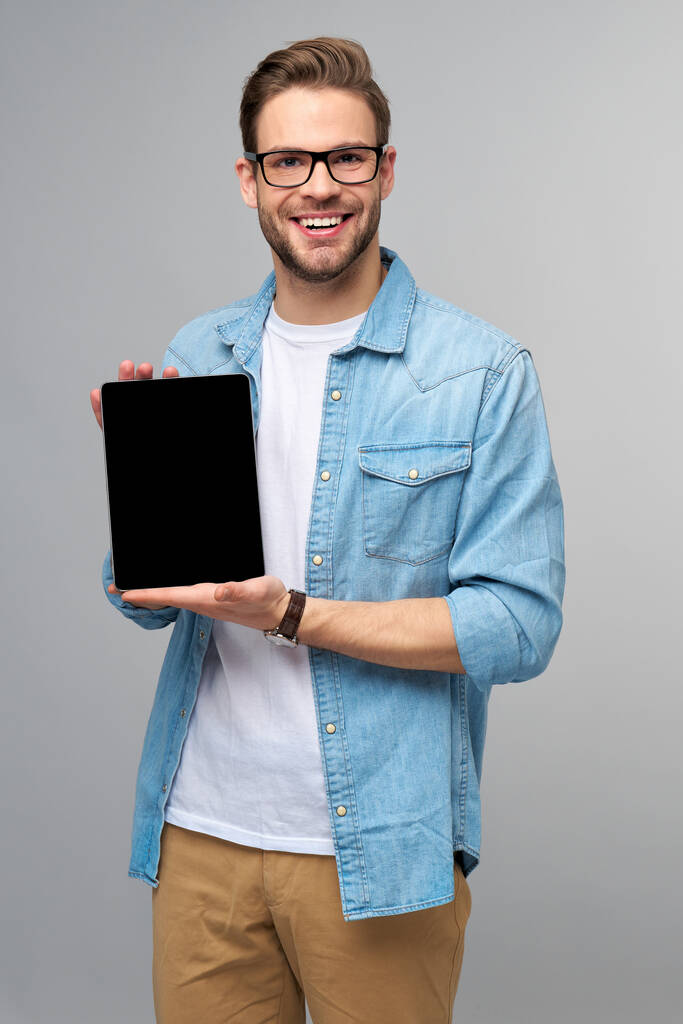 Jovem feliz vestindo camisa jeans de pé e segurando tablet pc pad sobre estúdio fundo cinza - Foto, Imagem