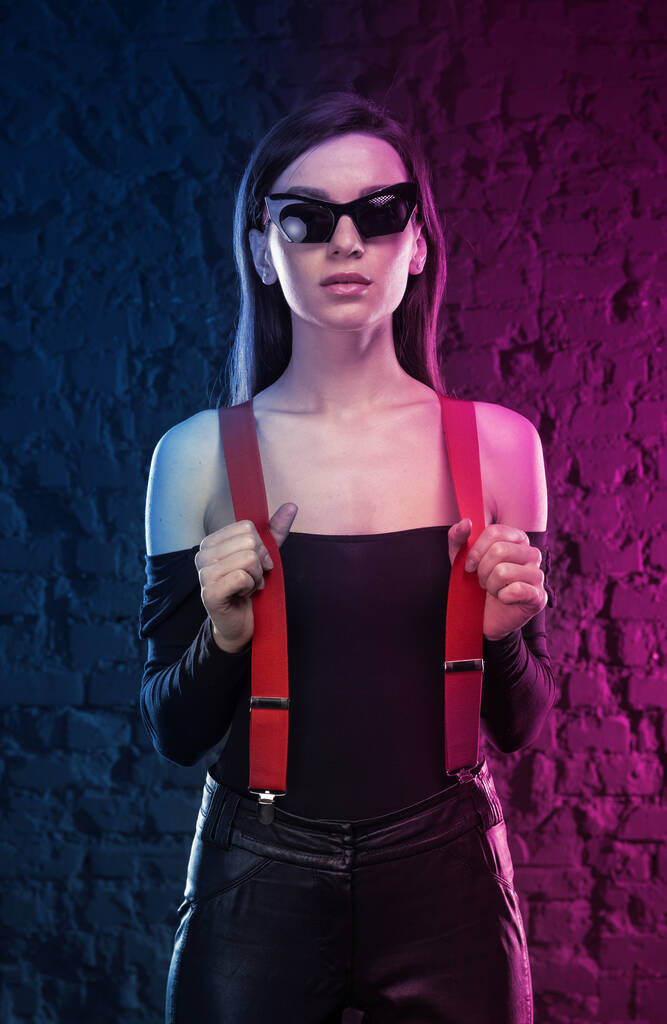 Mulher elegante cyberpunk, rosa néon e luz de fundo azul. Menina na moda em óculos escuros e calças de couro. Estilo futurista. - Foto, Imagem