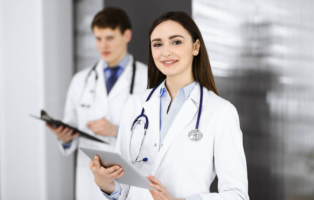 Hymyilevä älykäs naislääkäri pitää kädessään tablettitietokonetta, kun hän seisoo taustalla kollegansa kanssa klinikalla. Lääkärin muotokuva työssä. Täydellinen lääketieteellinen palvelu - Valokuva, kuva