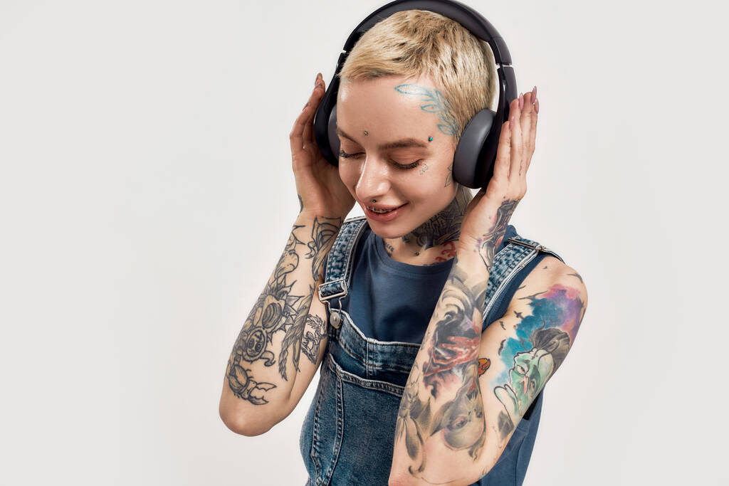 Listening to music. A young white pierced and tattoed woman with her eyes closed and hands over her ears wearing a denim overall listening to music with her big earphones - Photo, Image