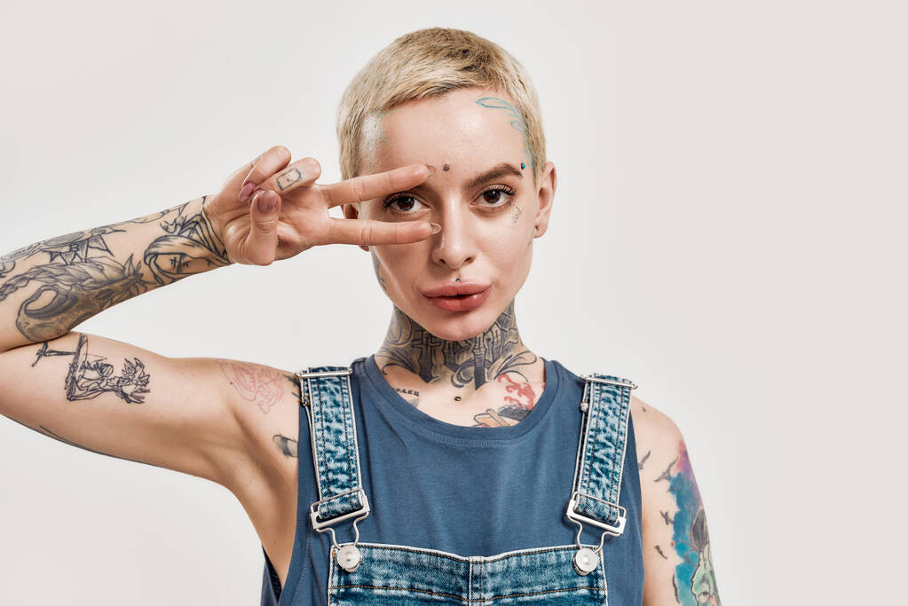 Эмоции и позы. Белая женщина с пирсингом и татуировкой, позирующая перед глазом, глядя в камеру в джинсе. - Фото, изображение