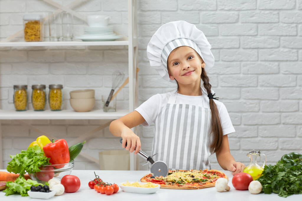roztomilá holčička v čepici šéfkuchaře a zástěra krájení pizzy - Fotografie, Obrázek