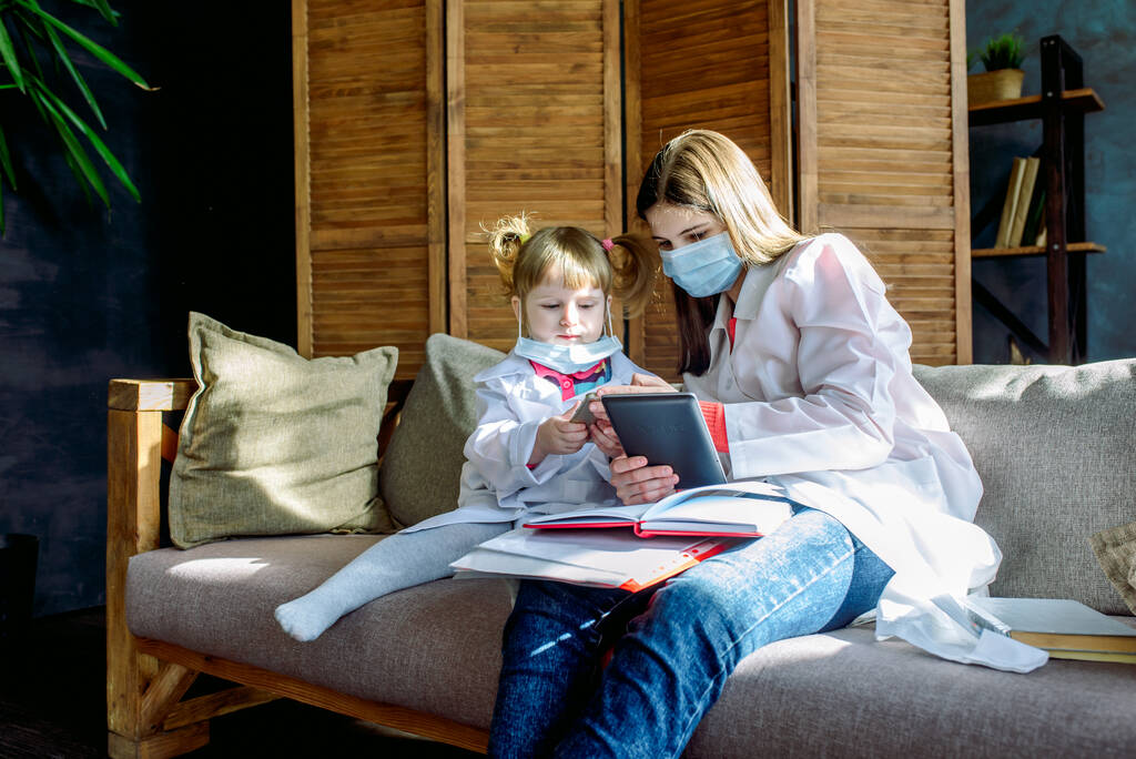 Dvě roztomilé sestřičky si doma na gauči hrají na doktorku. Lékařská uniforma, respirační maska, ochrana proti koronaviru. COVID-19 - Fotografie, Obrázek