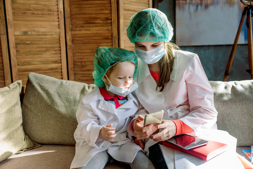 Two cute children play doctor and hospital using stethoscope. girl in uniform of a doctor and in face mask. Coronavirus concept, covid-19.Friends girls having fun at home or preschool. - Photo, Image