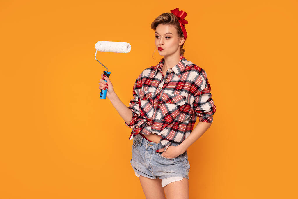 Female decorator painting wall indoors. Home repair service. A lot of copy space. Orange studio background. Girl in checkered shirt and short jeans. - Photo, Image