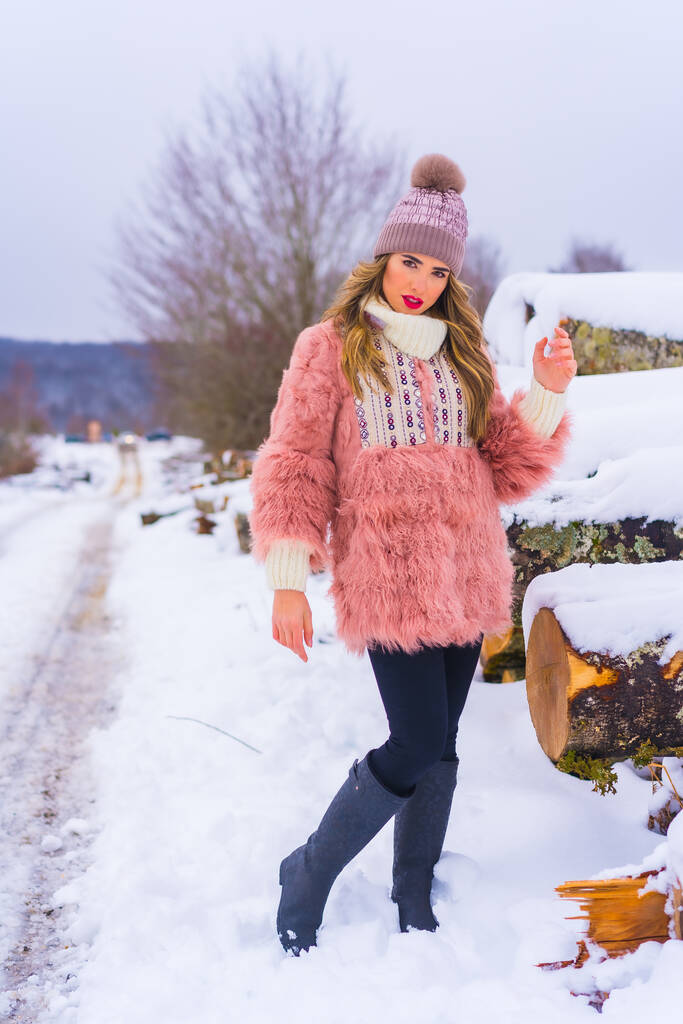 Modello biondo in posa con giacca di pelliccia rosa e cappello viola sulla neve. Accanto ad alcuni alberi tagliati sul sentiero, stile di vita invernale - Foto, immagini