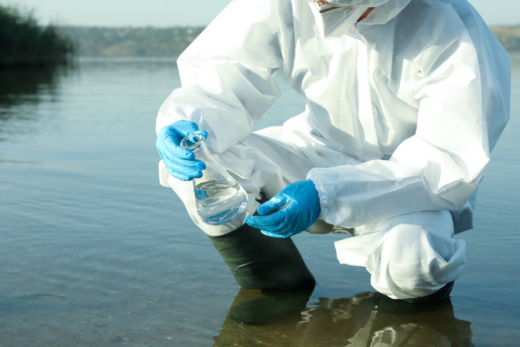 Kemiallisessa suojapuvussa oleva tutkija, jolla on erlenmeyerpullo ja joka ottaa näytteen joesta analysoitavaksi, lähikuva - Valokuva, kuva