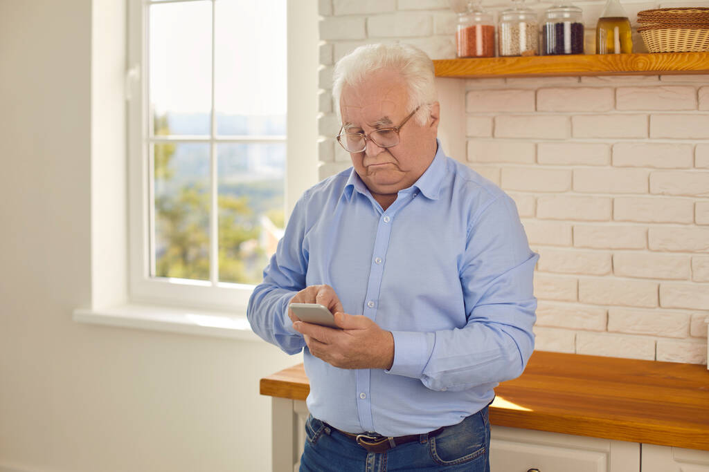 Usta adam akıllı telefona bakar ve evde mutfakta dikilirken haberleri dikkatle okur.. - Fotoğraf, Görsel