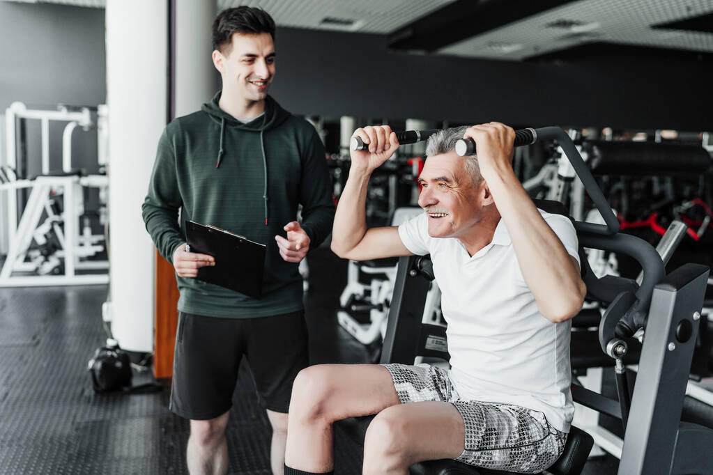 Fitness entrenador lleva a cabo la rehabilitación en el gimnasio con un hombre sinior, sonríe y le dice cómo realizar ejercicios. - Foto, imagen
