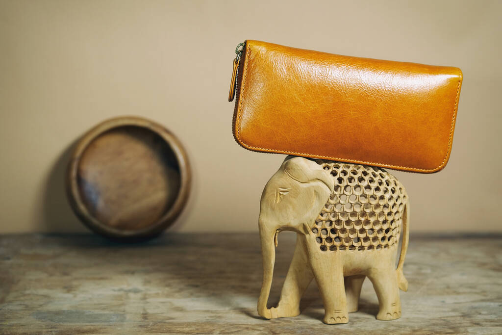 Ginger skórzana torebka leży z tyłu drewnianego słonia. Akcesoria modowe w indyjskim stylu folklorystycznym. Skład w kolorach natury, ton ziemi. - Zdjęcie, obraz