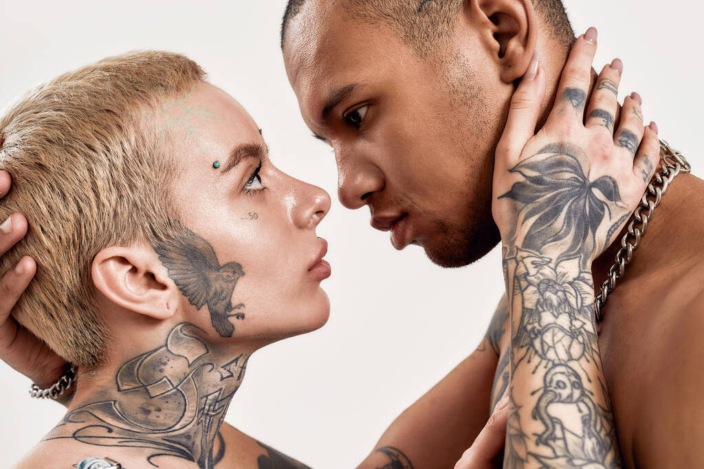 Мультикультурные отношения. Мужчина с топлесс-темнокожей татуировкой и белая женщина с пирсингом и татуировками, смотрящая друг другу в глаза, обнимаясь - Фото, изображение