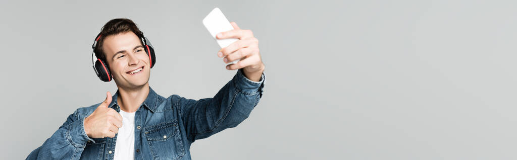 Gülümseyen adam, akıllı telefonuyla selfie çekerken gri, afişte izole edilmiş kulaklıklar kullanıyor. - Fotoğraf, Görsel