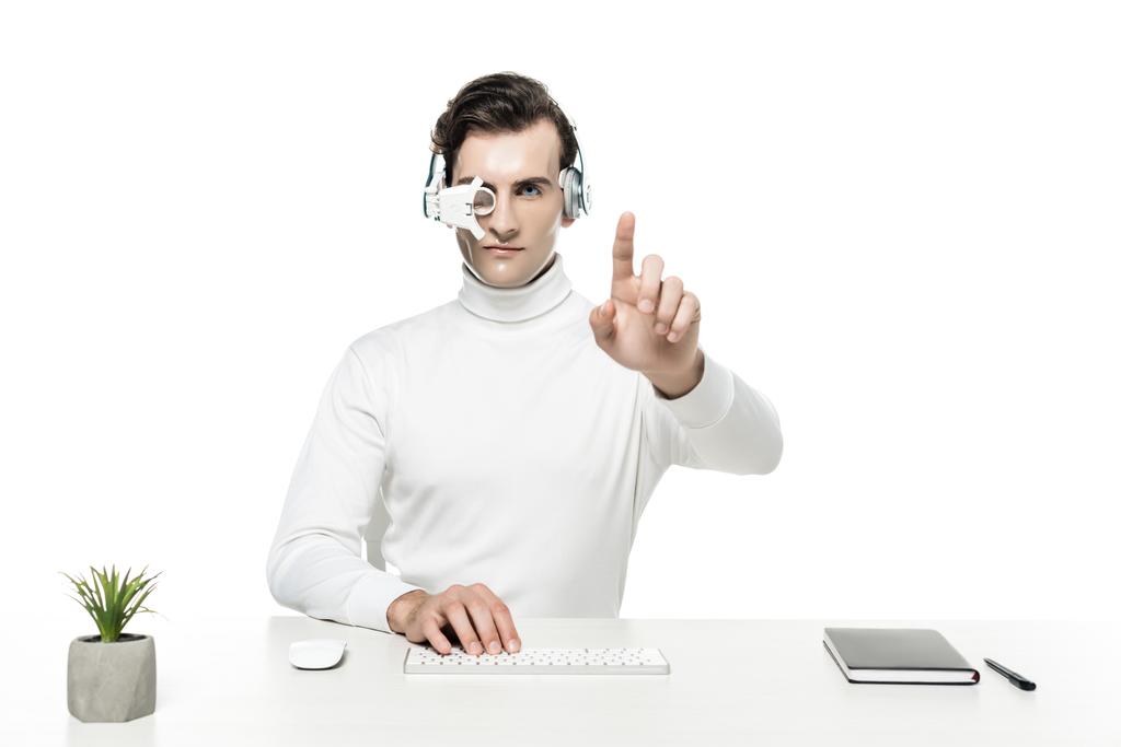 Киборг в наушниках и линзах для глаз, трогающий что-то и использующий клавиатуру компьютера возле ноутбука и растения, изолированного на белом - Фото, изображение