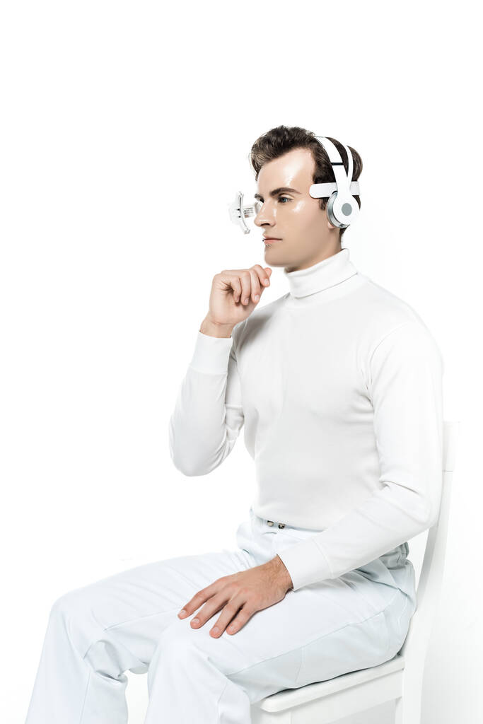 Cyborg hombre en lente para los ojos y auriculares con la mano cerca de la barbilla sentado en la silla aislada en blanco - Foto, Imagen