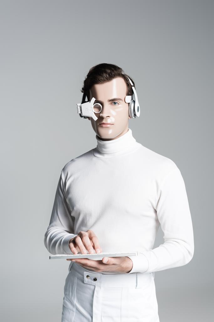 Cyborg en auriculares y lente digital que sostiene el teclado de la computadora y mira a la cámara aislada en gris - Foto, Imagen