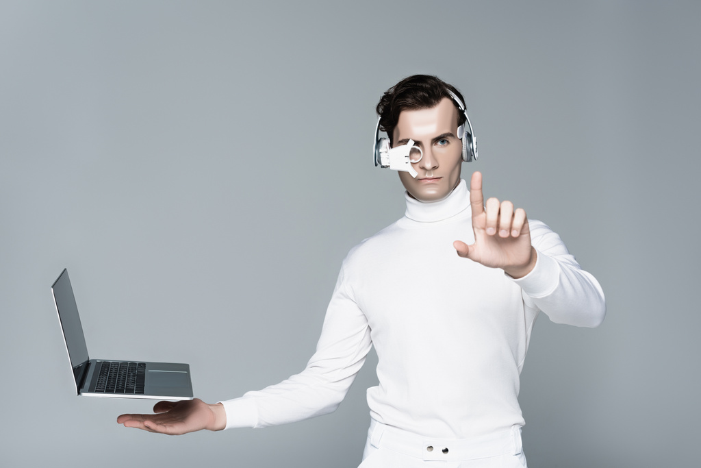 Cyborg in headphones touching something near laptop with blank screen levitating in air isolated on grey - Photo, Image