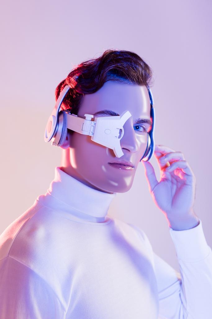 Cyborg mit Kopfhörer und Augenlinse blickt auf Kamera auf lila Hintergrund - Foto, Bild