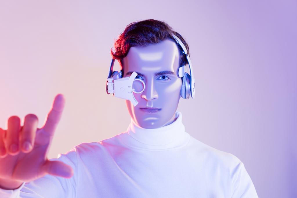 Cyborg σε ψηφιακό φακό ματιών και ακουστικά που δείχνουν με το δάχτυλο σε θολή πρώτο πλάνο σε μωβ φόντο - Φωτογραφία, εικόνα