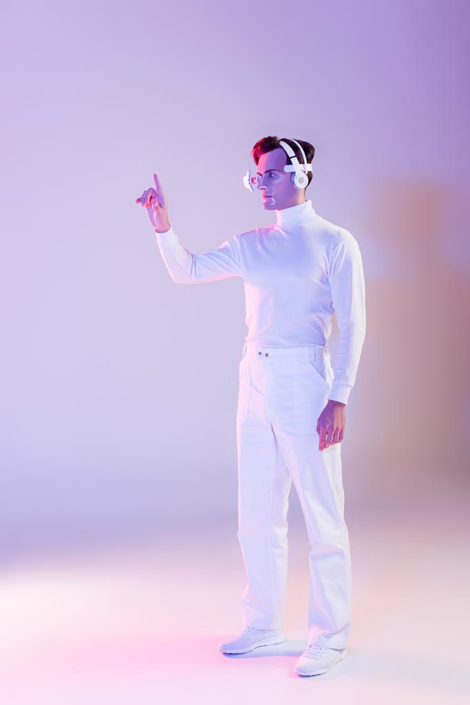 Cyborg en ropa blanca, lentes para los ojos y auriculares apuntando con el dedo hacia arriba sobre fondo púrpura - Foto, Imagen