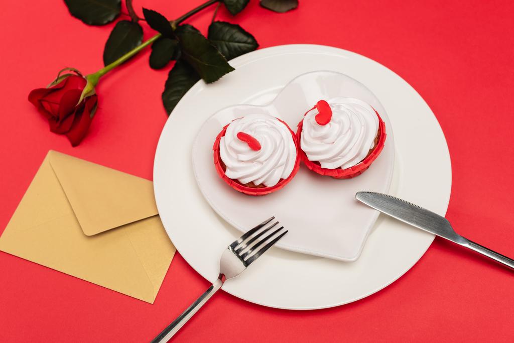 cupcakes op plaat in de buurt van roos en envelop op rode achtergrond - Foto, afbeelding