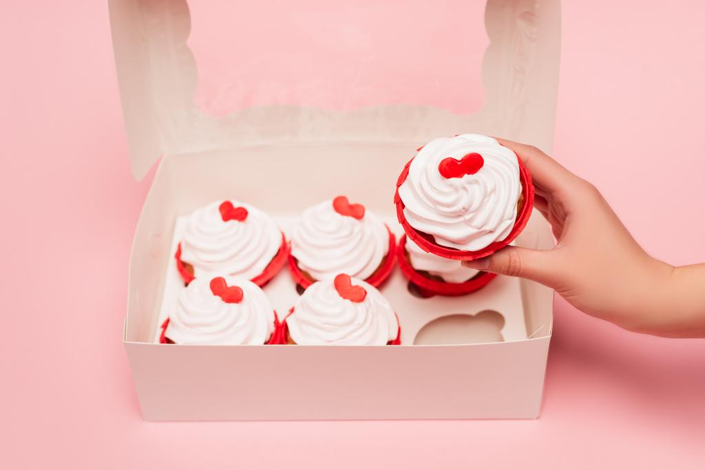 ピンクの背景の箱の近くにバレンタインカップケーキを持っている女性のクロップドビュー - 写真・画像