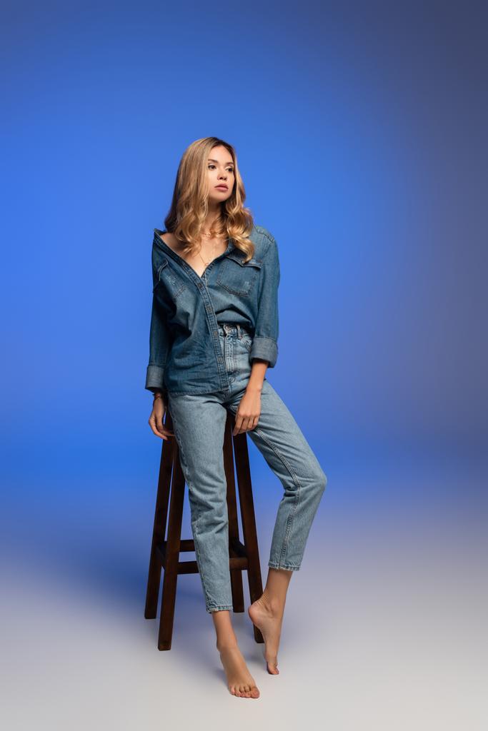 sensuele blootsvoets vrouw in stijlvolle denim kleding weg te kijken in de buurt van hoge stoel op blauw - Foto, afbeelding