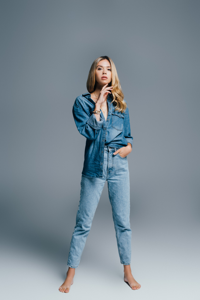 stylish, barefoot woman in denim shirt and jeans looking at camera while posing on grey - Photo, Image