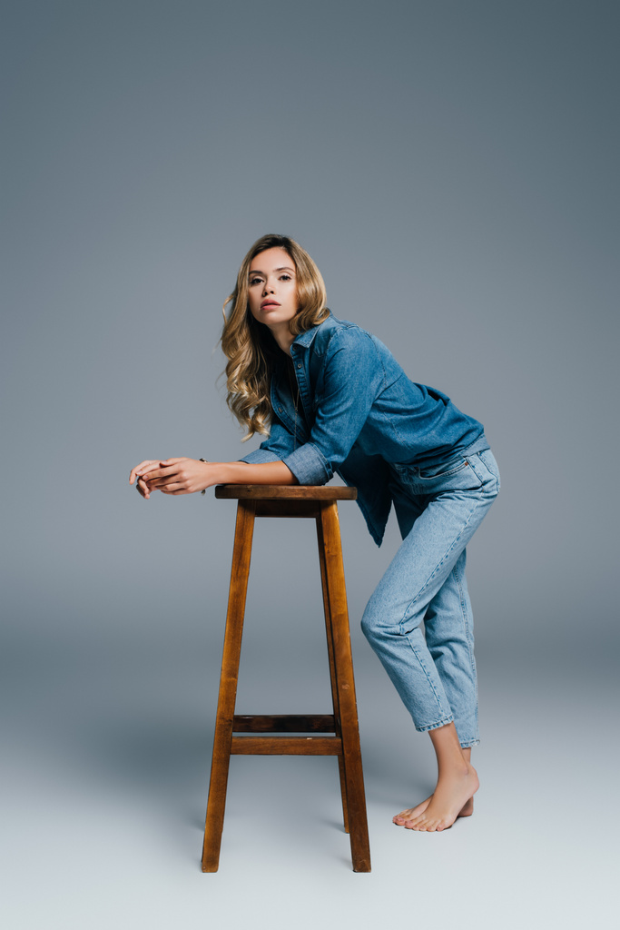 sensual woman in denim shirt and jeans leaning on high stool on grey - Photo, Image