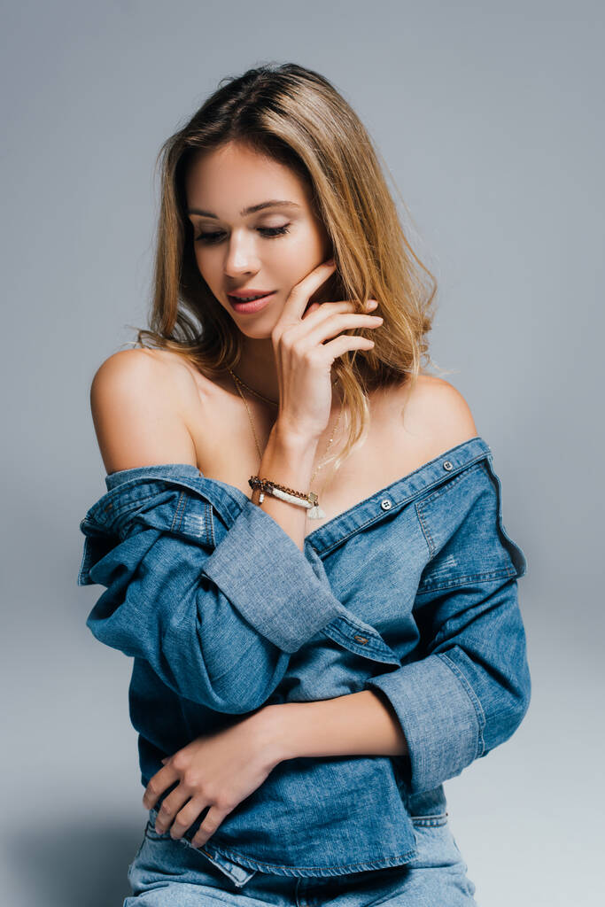seductive young woman in unbuttoned denim shirt, with naked shoulders, touching face  on grey - Photo, Image
