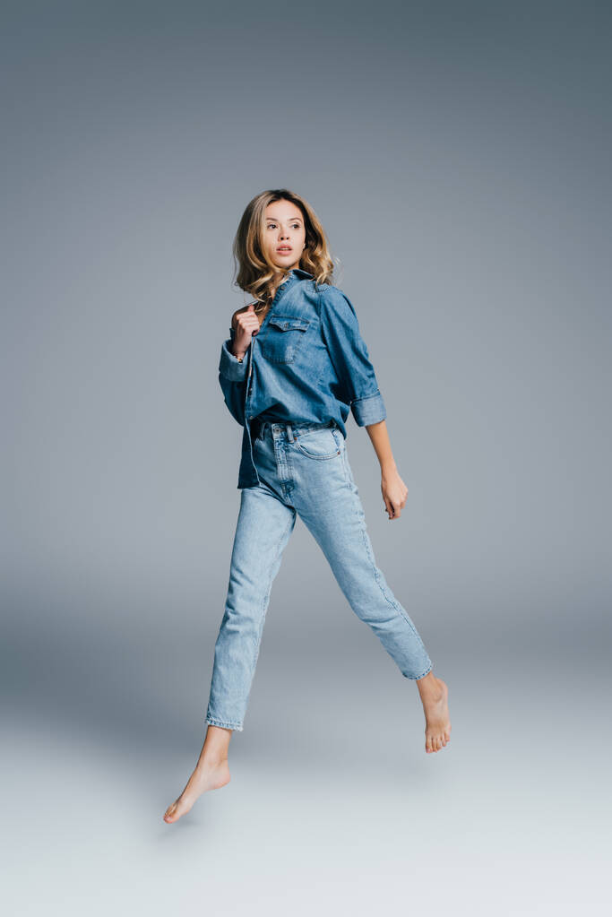 young barefoot woman in denim shirt and jeans looking away while levitating on grey - Photo, Image