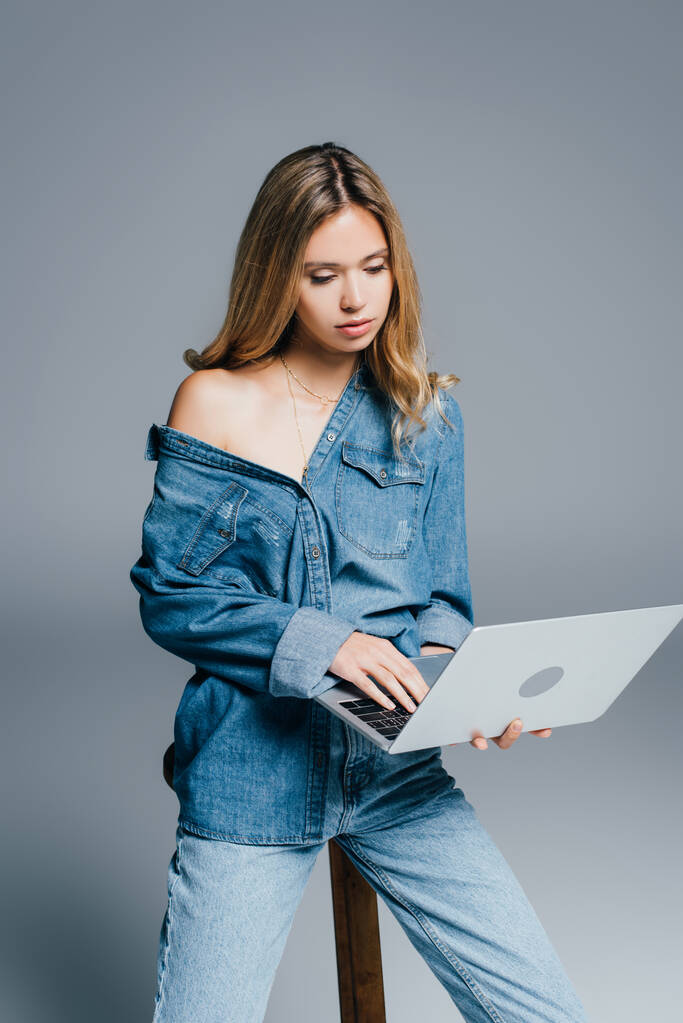 sinnliche junge Frau in Jeanskleidung, mit nackter Schulter, mit Laptop auf grau - Foto, Bild