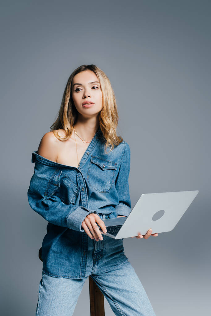 стильна жінка в джинсовій сорочці, з голим плечем, дивиться вбік, тримаючи ноутбук на сірому
 - Фото, зображення